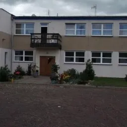 Dwupiętrowy budynek z balkonem nad wejściem, otoczony zielenią i utwardzonym terenem.