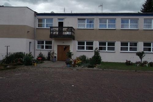 Dwupiętrowy budynek z balkonem nad wejściem, otoczony zielenią i utwardzonym terenem.