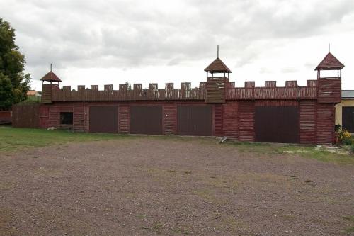 Drewniana konstrukcja garażowa stylizowana na średniowieczny zamek z wieżyczkami i blankami, z trzema bramami garażowymi, na żwirowym podłożu.