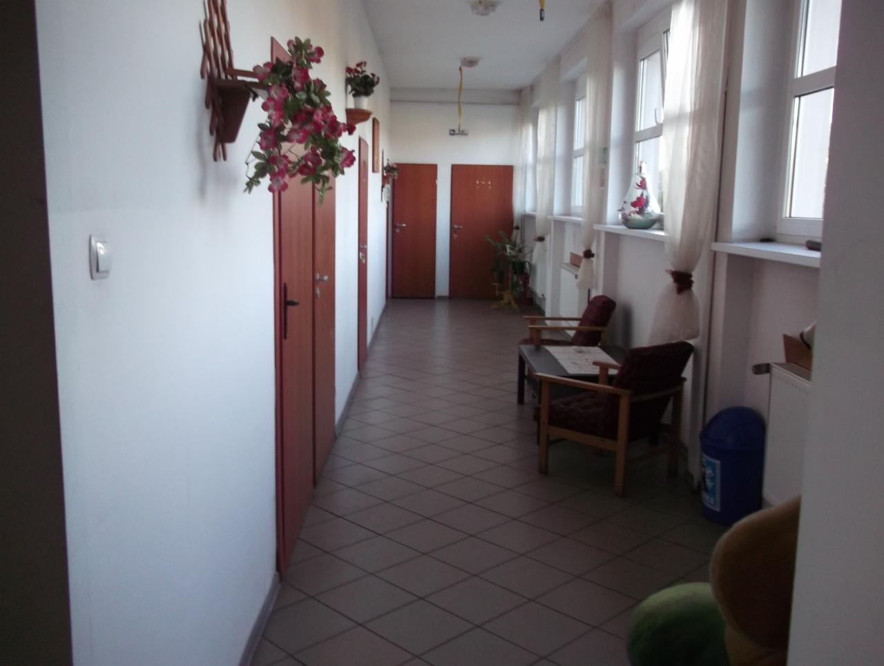 Korytarz budynku z brązowymi drzwiami, kwiatami na ścianach, oknami z białymi firankami i miejscem do siedzenia z dwoma krzesłami i stolikiem.