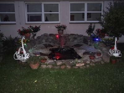 Ogródek z oczkiem wodnym otoczonym kamieniami i dekoracjami, w tle budynek z oknami.