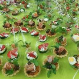 Różnorodne przekąski na zielonym obrusie: tartaletki z pastą i szynką parmeńską, ogórki z serkiem i truskawkami, sałatki w przezroczystych naczyniach.