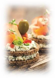 Dwa apetyczne, wielowarstwowe kanapeczki na ciemnym pieczywie, udekorowane kremowym serem, łososiem, papryką, oliwką i świeżymi ziołami, ułożone na drewnianej desce, z rozmytym tłem.