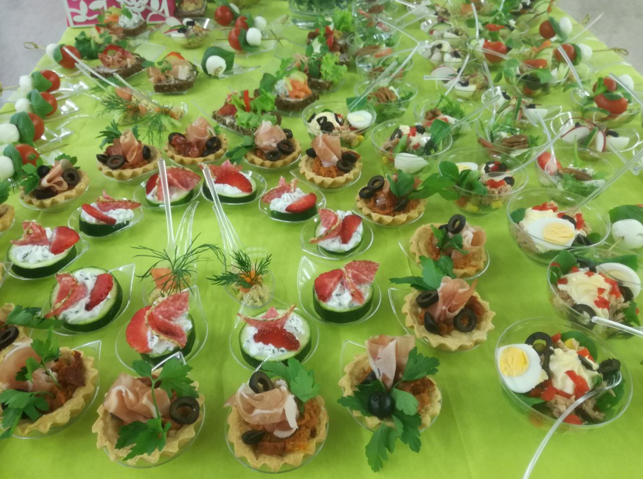 Różnorodne przekąski na zielonym obrusie: tartaletki z pastą i szynką parmeńską, ogórki z serkiem i truskawkami, sałatki w przezroczystych naczyniach.