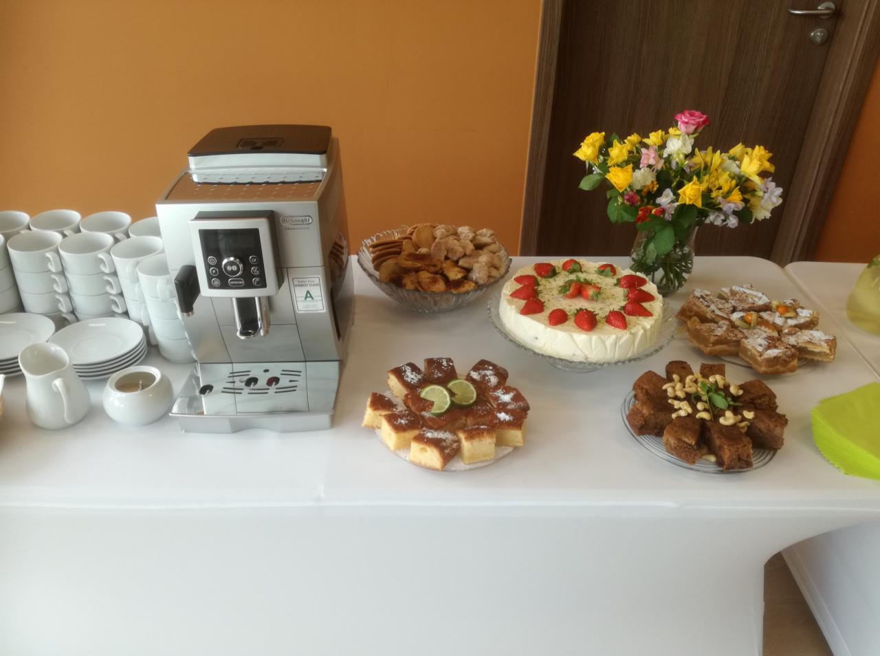 Stół z białym obrusem zastawiony różnorodnymi ciastami, ciasteczkami, deserami, kawą z ekspresu Delonghi i bukietem kwiatów w szklanym wazonie, gotowy na przyjęcie.