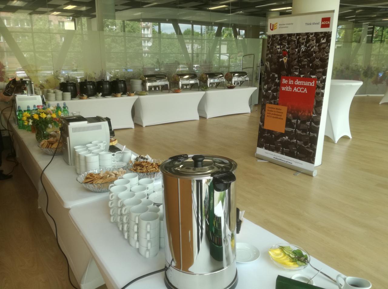 Stół z cateringiem na konferencji, z automatami do kawy, ciastkami, owocami i napojami, za stołem widoczny baner reklamowy.