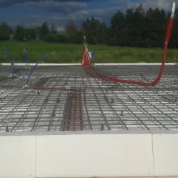 Przygotowanie płyty fundamentowej z izolacją termiczną, zbrojeniem i instalacją hydrauliczną; widoczne niebieskie i czerwone rury oraz siatka zbrojeniowa.