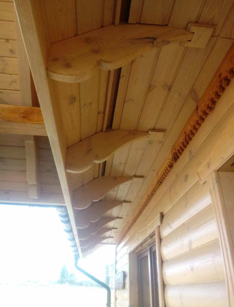 Drewniany okap domu z widocznymi zdobieniami i rynnami, wykonany z jasnego drewna, z dekoracyjnymi wspornikami i ornamentami wzdłuż krawędzi dachu.