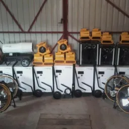 Różne urządzenia marki Trotec do osuszania, w tym wentylatory podłogowe, nagrzewnice i osuszacze kondensacyjne, ustawione rzędami w magazynie z blachy falistej.