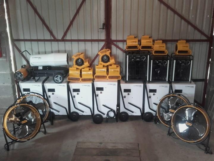 Różne urządzenia marki Trotec do osuszania, w tym wentylatory podłogowe, nagrzewnice i osuszacze kondensacyjne, ustawione rzędami w magazynie z blachy falistej.