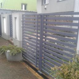 FHU ART-MET Rafał Fórmaniak - Nowoczesna, szara brama wjazdowa z poziomymi panelami, betonowe słupki z dekoracyjnymi wnękami, kostka brukowa i roślinność w tle.