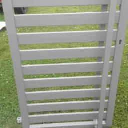 Szara, metalowa furtka z poziomymi szczebelkami, widok z bliska na trawniku, z elementami konstrukcyjnymi widocznymi w tle.