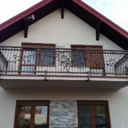 Kuta balustrada balkonowa z motywem winorośli na tle białej elewacji domu z brązowym dachem.