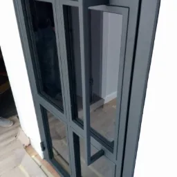 Szklane drzwi wewnętrzne z metalową ramą w kolorze grafitowym i prostokątnym uchwytem, widoczne zabezpieczenia podłogi podczas montażu.