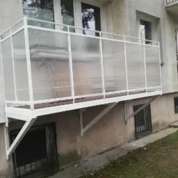 Zabudowany balkon z białą ramą i przezroczystymi panelami z tworzywa sztucznego, wsparty na wspornikach przymocowanych do ściany budynku.