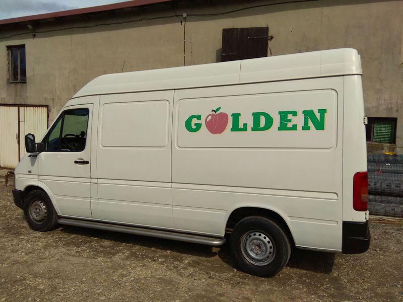 Biały samochód dostawczy z zielonym napisem 'GOLDEN' i grafiką jabłka na boku, stojący na szutrowej nawierzchni przed budynkiem gospodarczym.