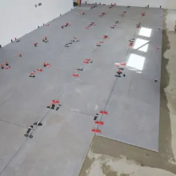 Szare płytki podłogowe w trakcie układania, z widocznymi systemami poziomowania płytek w kolorze czerwonym i czarnym, poziomica oparta o ścianę w tle.