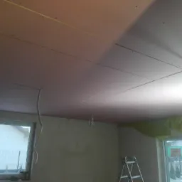 Montaż płyt kartonowo-gipsowych na suficie, widoczne profile, przewody elektryczne i drabina w pomieszczeniu z oknami.