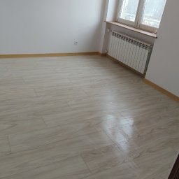 Rad-Bud - Jasne panele podłogowe w nowym, pustym pokoju z białymi ścianami i grzejnikiem pod oknem. Listwy przypodłogowe w kolorze drewna. Widoczny fragment futryny.