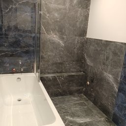 Rad-Bud - Wykończona łazienka z wanną obudowaną płytkami imitującymi marmur, szklany parawan nawannowy, płytki podłogowe i ścienne w tym samym stylu, oświetlenie sufitowe.