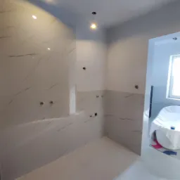 Świeżo wykafelkowana łazienka z marmurowymi płytkami, widoczne otwory na armaturę, przygotowana do dalszych prac wykończeniowych, zabezpieczona wanna w tle.