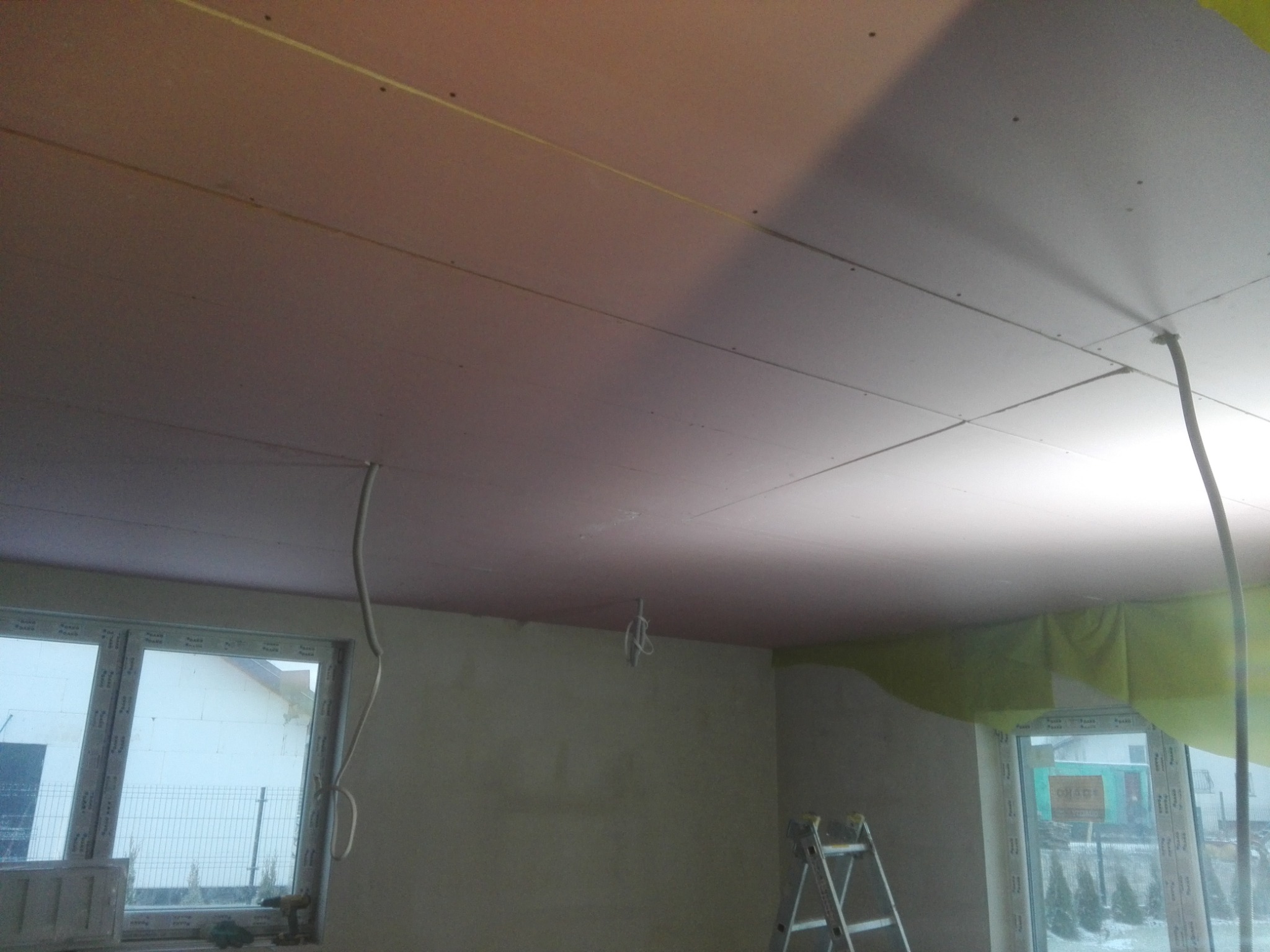 Montaż płyt kartonowo-gipsowych na suficie, widoczne profile, przewody elektryczne i drabina w pomieszczeniu z oknami.