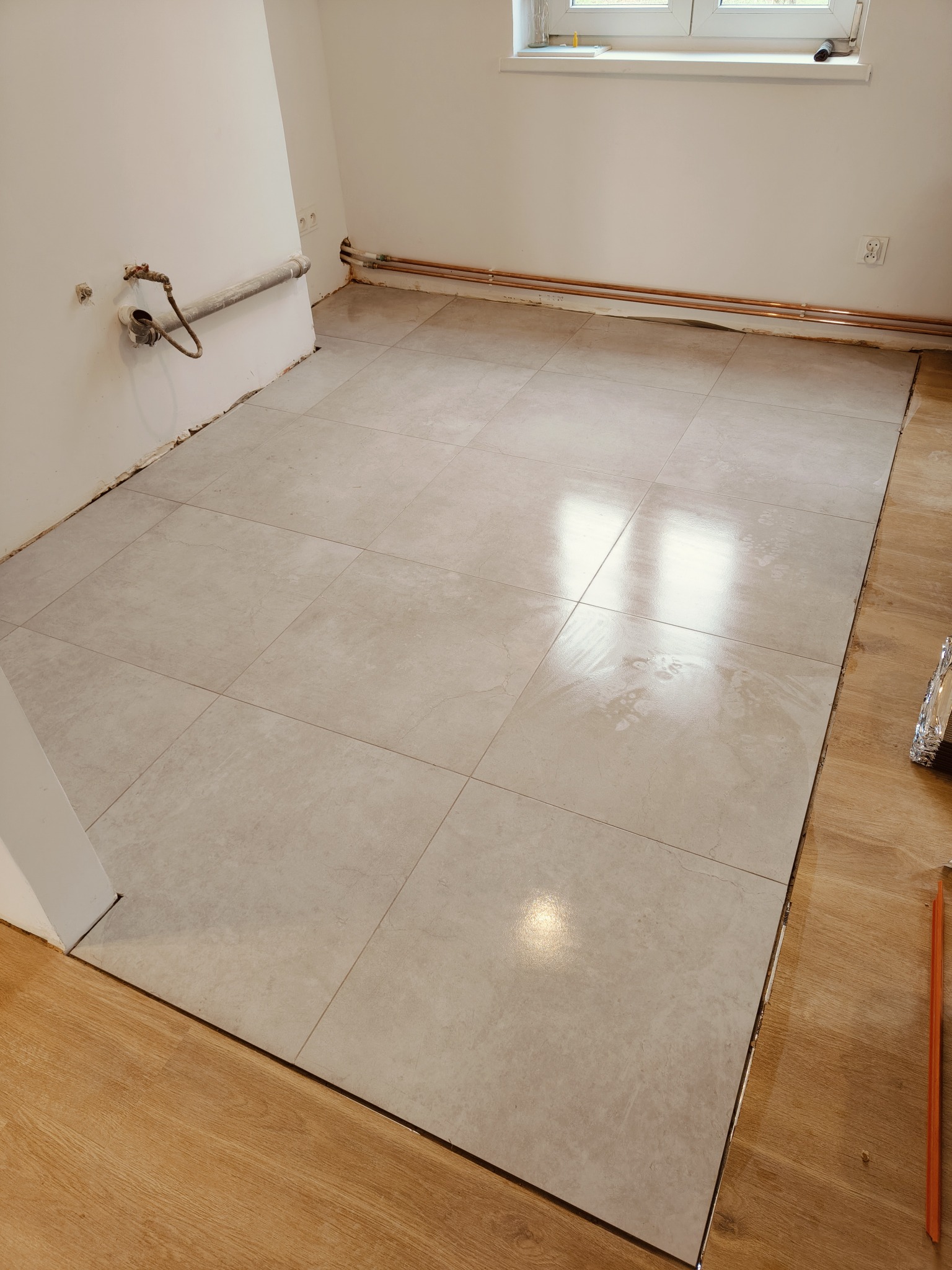 Ułożona podłoga z jasnych, kwadratowych płytek w pomieszczeniu z instalacją hydrauliczną przy ścianie i fragmentami drewnianej podłogi. Widoczne rury miedziane wzdłuż ściany.