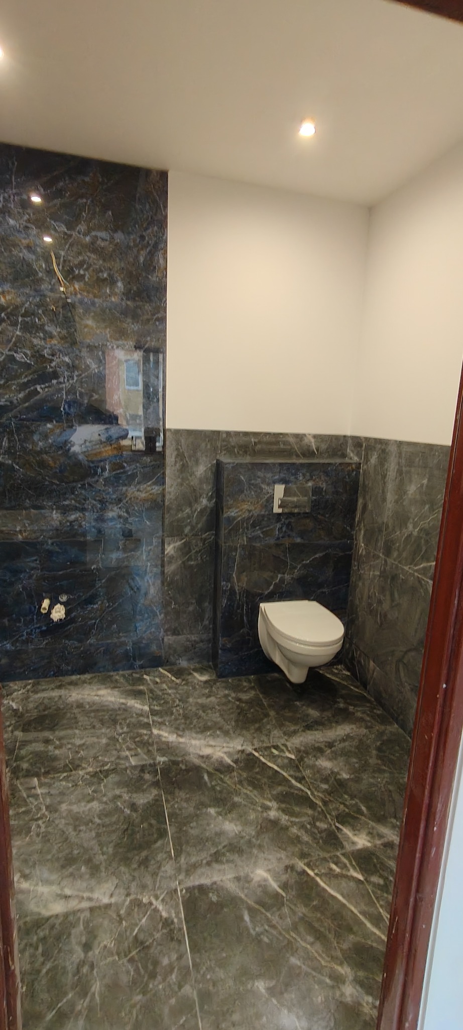 Wykończona łazienka z marmurowymi płytkami w odcieniach szarości i bieli. Wisząca toaleta, minimalistyczny design, oświetlenie punktowe. Widok zza framugi drzwi.