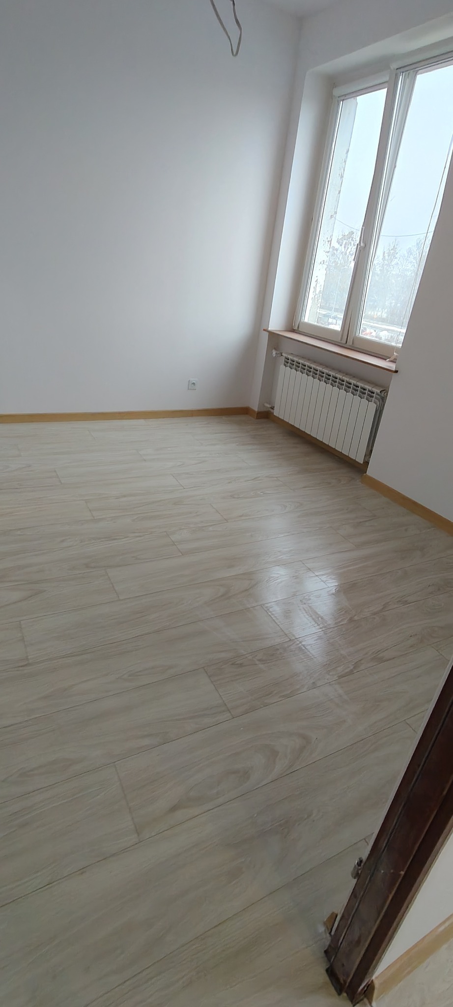 Jasne panele podłogowe w nowym, pustym pokoju z białymi ścianami i grzejnikiem pod oknem. Listwy przypodłogowe w kolorze drewna. Widoczny fragment futryny.