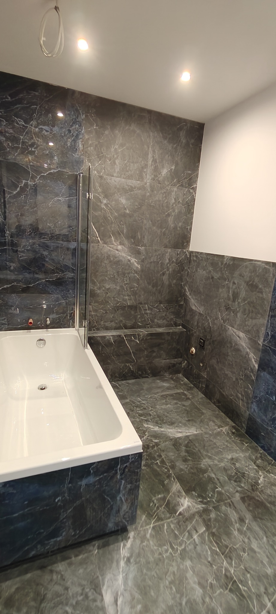 Wykończona łazienka z wanną obudowaną płytkami imitującymi marmur, szklany parawan nawannowy, płytki podłogowe i ścienne w tym samym stylu, oświetlenie sufitowe.
