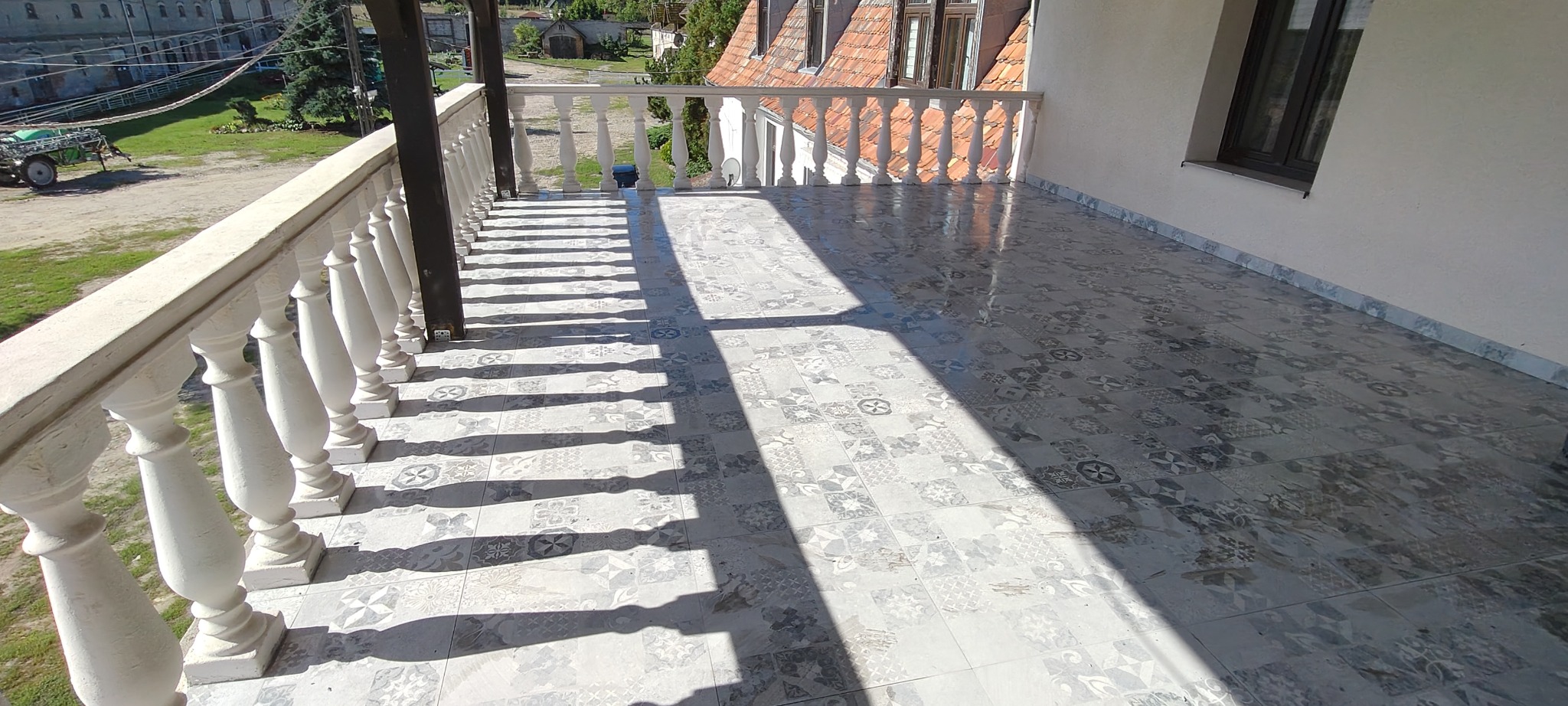 Wykończony balkon z balustradą i wzorzystą, szarą podłogą z płytek. Widok na zewnątrz z fragmentem budynku w tle. Cień balustrady na podłodze.