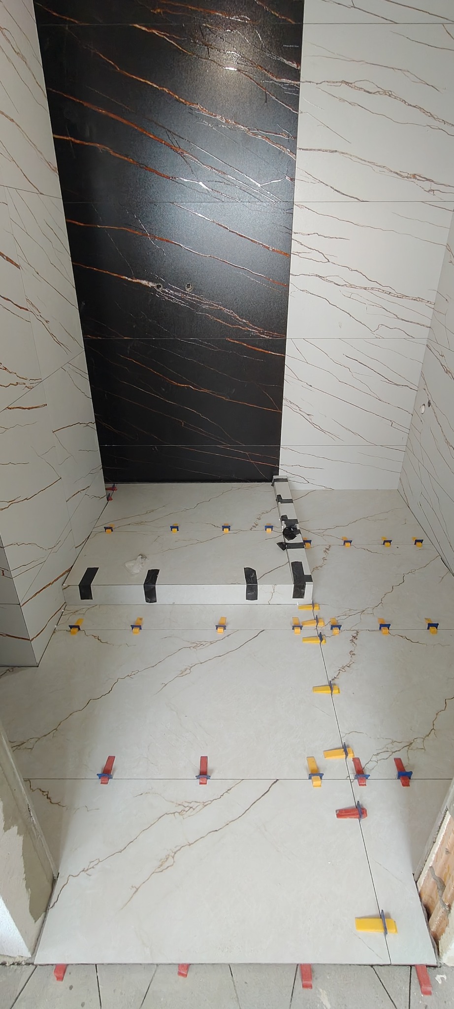 Układanie płytek imitujących marmur na podłodze i ścianach, z użyciem klipsów poziomujących. Widoczna czarna ściana z brązowymi żyłami. Ujęcie z góry.