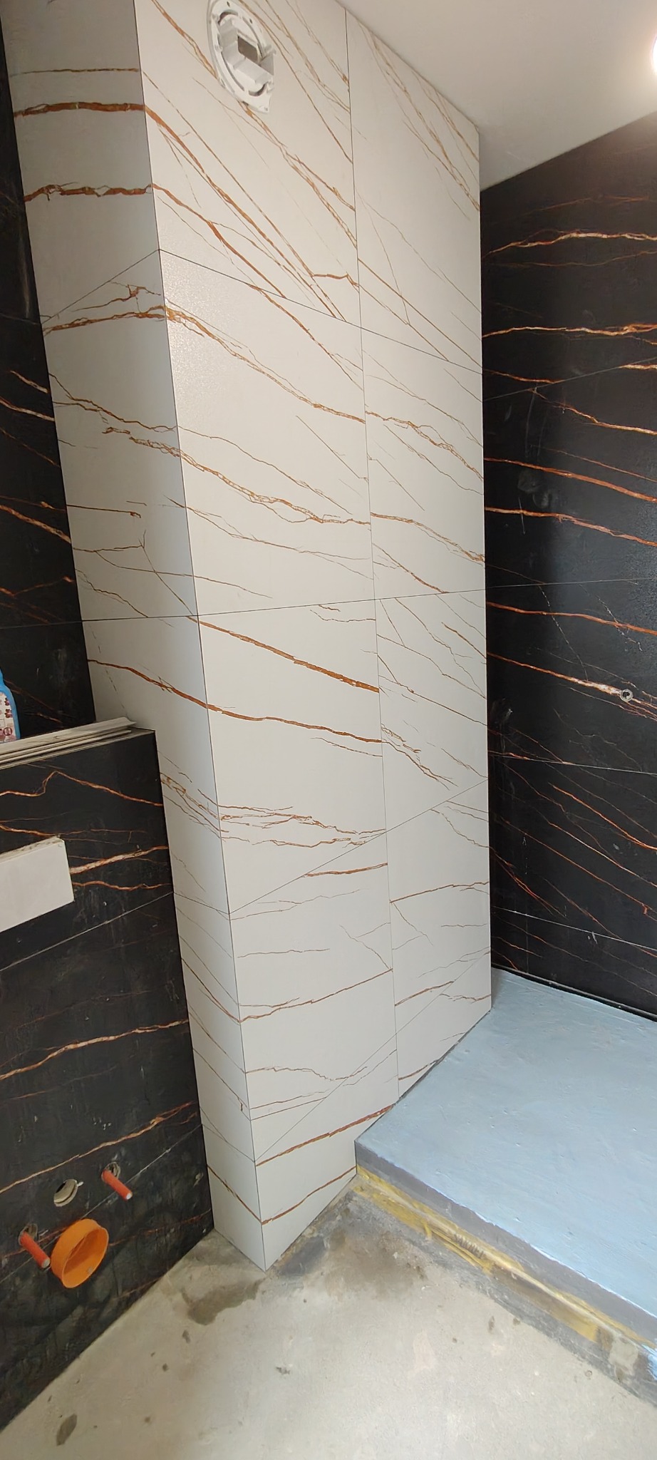 Wykończona kabina prysznicowa z białymi płytkami imitującymi marmur z brązowymi żyłami, kontrastująca z ciemną ścianą. Widoczne elementy instalacji.