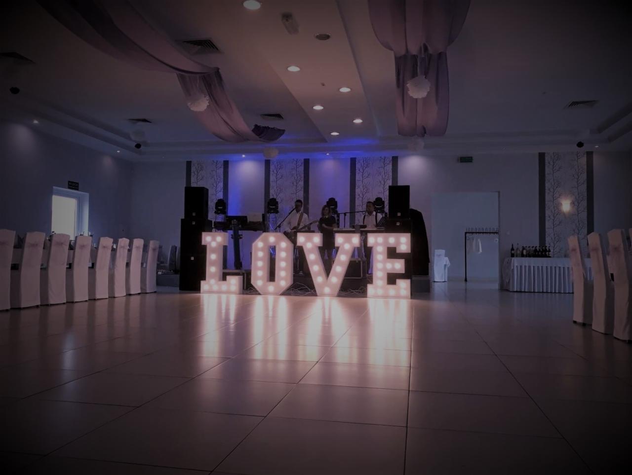 Sala bankietowa z dekoracją świetlną LOVE, zespołem muzycznym na scenie i przygotowanymi stołami z białymi pokrowcami na krzesłach.