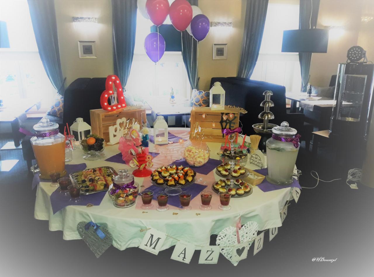 Stół z dekoracjami i przekąskami na przyjęcie, w tle balony, drewniane skrzynki z napisami i fontanna czekoladowa. Na stole napoje w szklanych słojach, owoce, słodycze i małe kanapeczki.