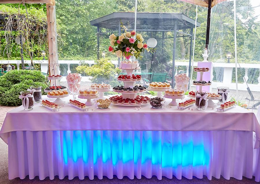 Stół z deserami na przyjęciu, oświetlony niebieskim światłem, z tortem na szczycie piramidy słodkości i bukietem róż w tle.