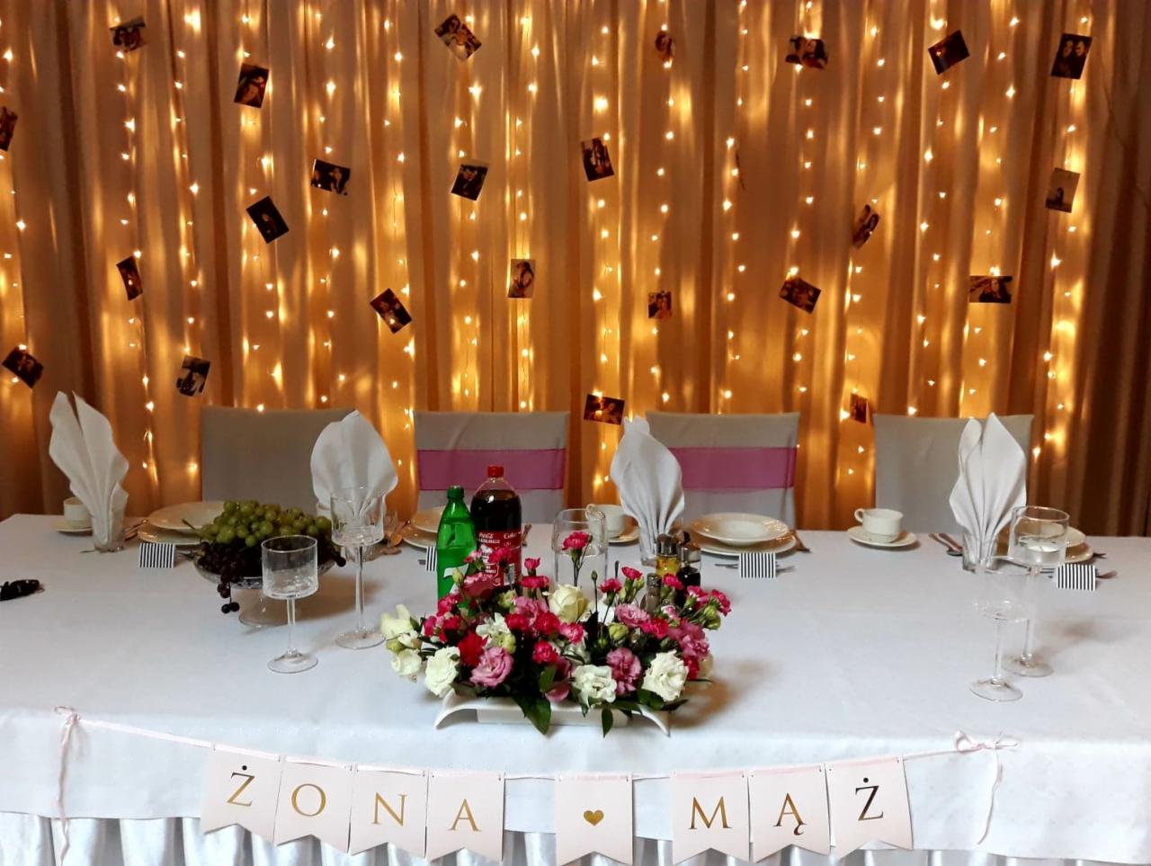 Stół weselny z białym obrusem, dekoracją kwiatową, owocami, napojami i girlandą z napisem 'Żona Mąż', tło z kurtyną świetlną i zdjęciami.