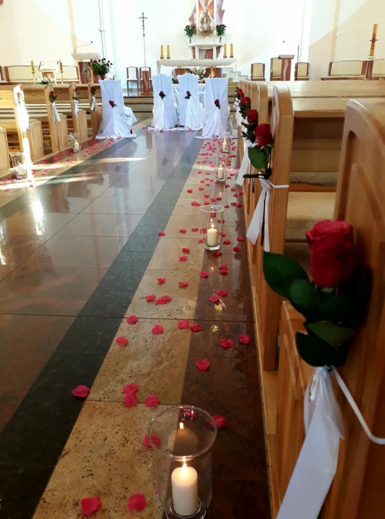 Uroczystość ślubna w kościele: przejście udekorowane płatkami róż i świecami w szklanych lampionach, ławki ozdobione różami i białymi wstążkami, krzesła w białych pokrowcach z różami.