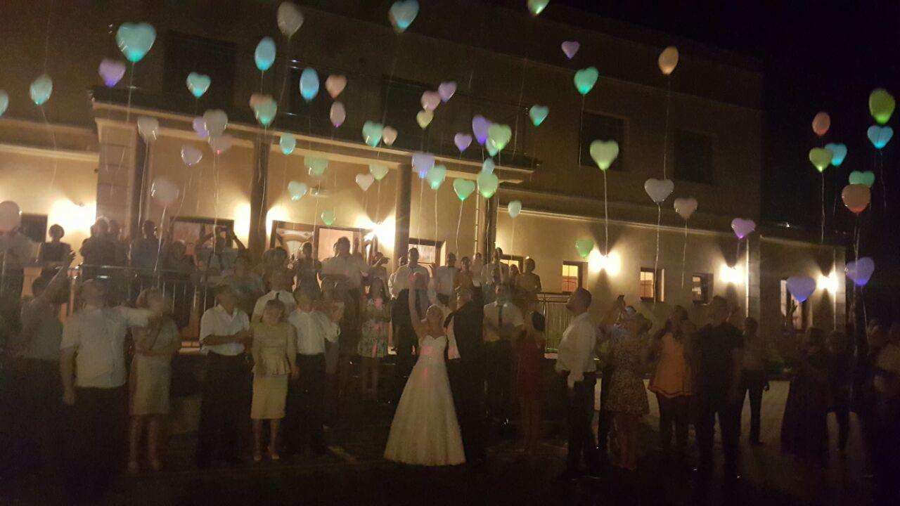 Wieczorne wypuszczanie w górę świecących balonów w kształcie serc przez parę młodą i gości weselnych przed budynkiem.