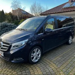 Granatowy van Mercedes V-klasy zaparkowany na brukowanym podjeździe przed domem z ogrodem. Widoczne detale: chromowane listwy, srebrne felgi i tablica rejestracyjna.