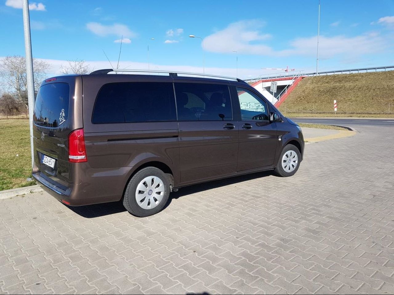 Brązowy Mercedes Vito z symbolem osoby niepełnosprawnej, zaparkowany w Warszawie na kostce brukowej, z widocznym wiaduktem w tle.