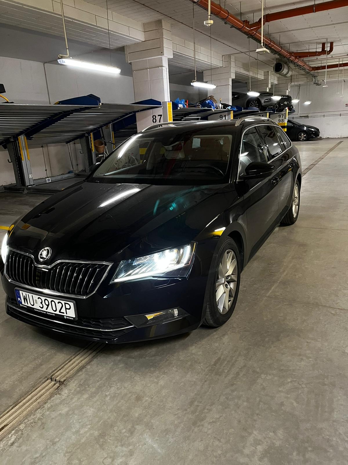 Czarna Skoda Superb Combi zaparkowana w garażu podziemnym, widoczne podnośniki samochodowe i inne pojazdy w tle.