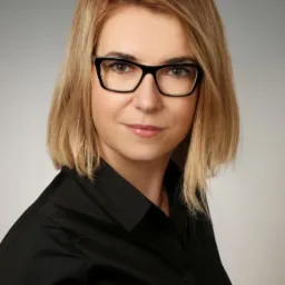 Portret kobiety z blond włosami w czarnej koszuli i okularach o grubych oprawkach, uśmiechająca się do kamery na neutralnym szarym tle.