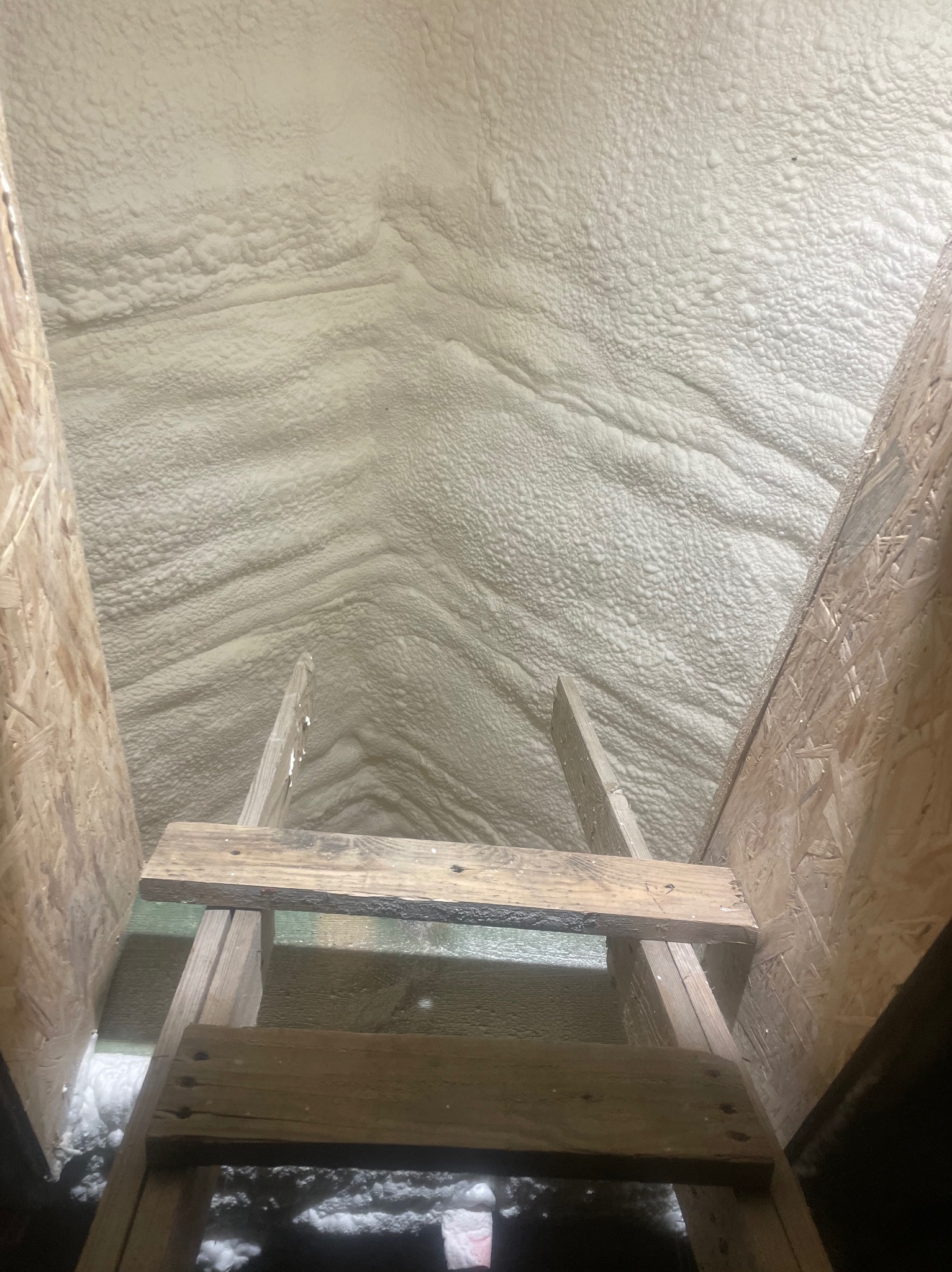 Strych po natrysku pianki PUR w Chojnie. Widok z drabiny na zaizolowany dach, widoczne drewniane elementy konstrukcji i biała, nierówna powierzchnia izolacji.