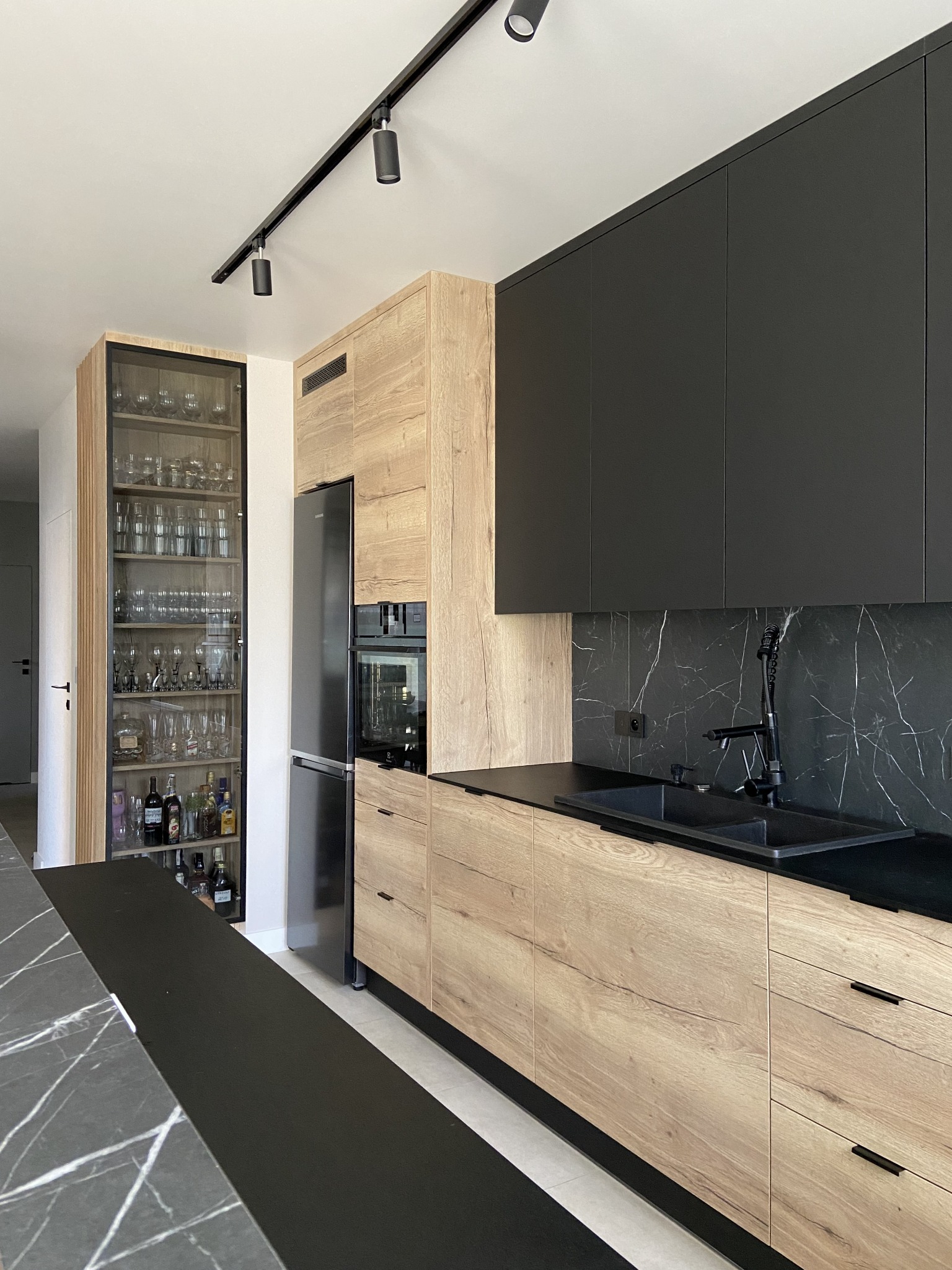 Nowoczesna kuchnia: czarne szafki wiszące, drewnopodobne szafki dolne, czarny blat z marmurowym wzorem, oświetlenie szynowe i przeszklona witryna z kolekcją szkła.
