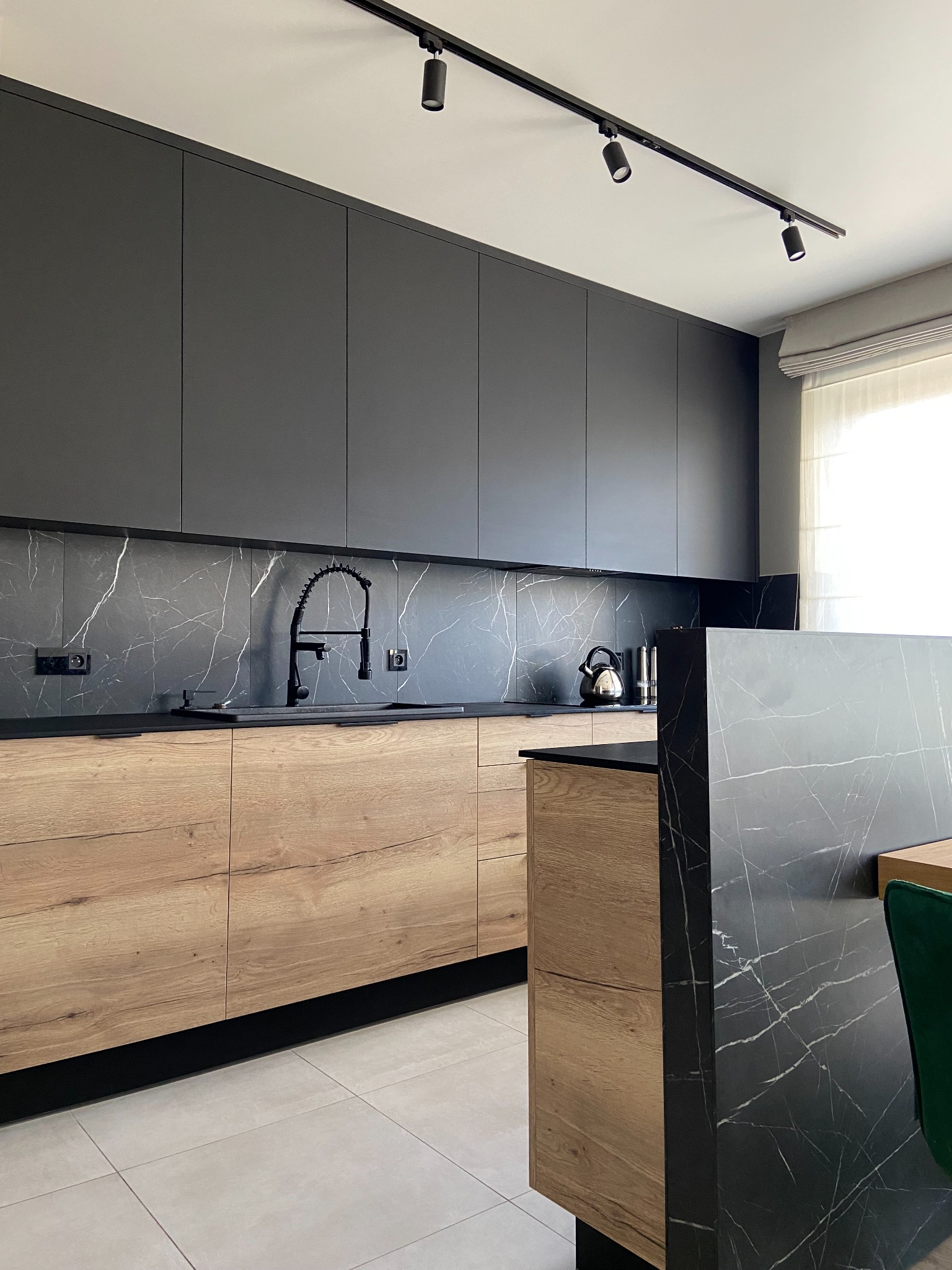 Nowoczesna kuchnia z czarnymi szafkami górnymi i drewnianymi dolnymi, marmurowy backsplash i wyspa, czarna armatura, oświetlenie szynowe.
