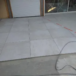 Układanie jasnych, kwadratowych płytek na zewnątrz budynku, widoczne krzyżyki dystansowe i przewód elektryczny na podłożu.