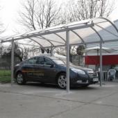 Zadaszenie samochodowe z przezroczystym poliwęglanem na metalowej konstrukcji, szary samochód osobowy zaparkowany pod spodem, widoczne słupy nośne i łukowata forma dachu, w tle drzewa i budynek.