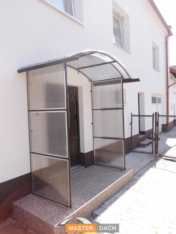 Zadaszenie wejścia do domu z poliwęglanu, wsparte na metalowej konstrukcji, widok z zewnątrz na jasny budynek z oknami.