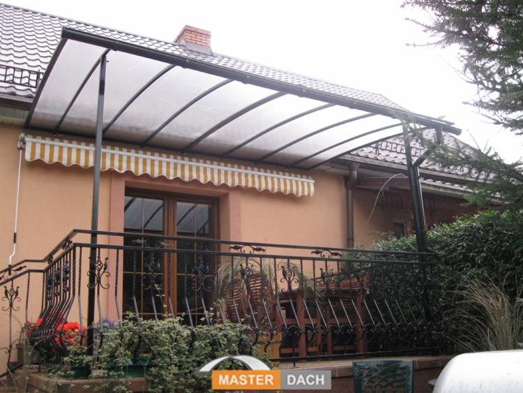 Zadaszony balkon z czarną, kutą balustradą, osłonięty przezroczystym daszkiem i markizą w biało-żółte pasy, z widocznymi roślinami w donicach i elementami ogrodu.